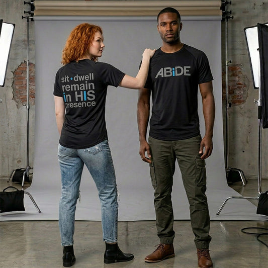 Two models wearing matching black ABiDE Luxury Bamboo t-shirts in a studio setting. The woman on the left shows the back of the shirt with the text 'sit • dwell remain in HIS presence' in Shadow Gray while the man shows the fron ABiDE logo in Shadow Gray & Electric Blue.