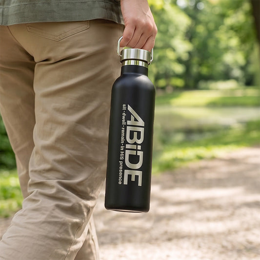A youth walking on a gravel trail in a treed area carrying a black Water Bottle with stainless steel screw on lid with handle, showing a laser engraved ABiDE sit - dwell - remain in HIS presence logo.