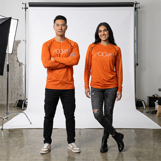 A man and a woman standing in front of a white studio backdrop, both wearing bright orange long-sleeve athletic shirts with a white 'ADONAI' logo on the chest, paired with dark pants.