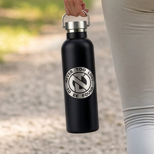 A zoomed in photo of a woman walking a pebble path in a park with grass in the background, carrying a Never Too Lost black Water Bottle with stainless steel screw on lid with handle showing a laser engraved signature Never Too Lost 'NTL' Logo with Never Too Lost To Be Found encircled around it.