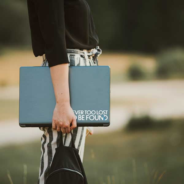 student standing in park with laptop in hand with Never Too Lost To Be Found sticker