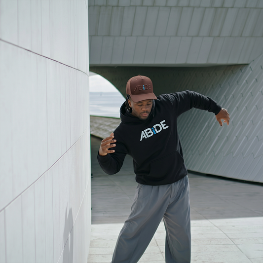 Man wearing a black ABiDE hoodie with 'ABIDE' text, gray pants, and Brown on Brown ABiDE Trucker hat in an urban setting.
