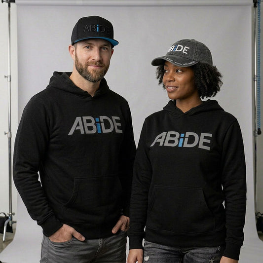 A man and a woman standing side by side in a studio setting, both wearing black pullover hoodies with the word 'ABiDE' printed across the chest in gray and blue, along with matching ABiDE branded caps.