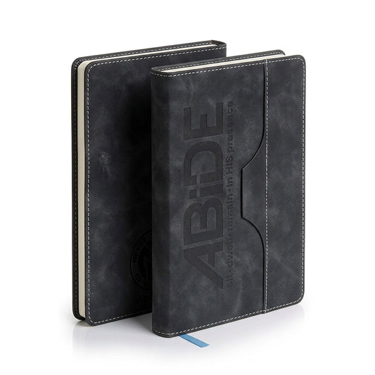 A black textured faux-leather ABiDE Journal standing upright, with a second matching notebook partially visible behind it. The front cover has debossed vertical branding reading 'ABIDE' with smaller text 'all around in HIS presence' and white contrast stitching around the edges.