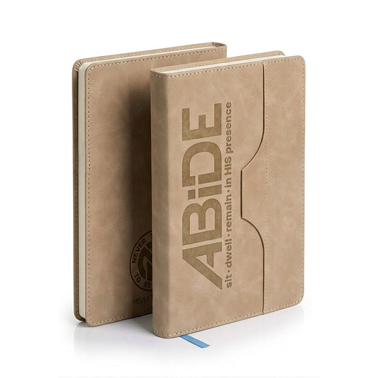 Two tan faux-leather journal covers standing upright on a white background. The front cover has large embossed text reading “ABiDE” with smaller text “sit · dwell · remain · in his presence,” and the back cover shows a circular NTL - Never Too Lost logo and a small blue ribbon bookmark.