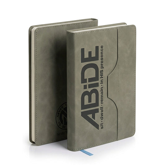 A gray faux-leather journal standing upright, with a second copy leaning behind it. The front cover features the text 'ABIDE' and 'sit - dwell - remain - in HIS presence' printed in dark gray and rear has cicular NTL - Never Too Lost Logo.