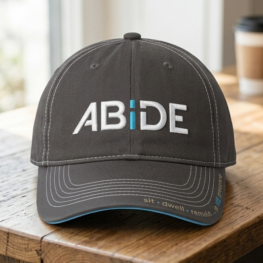 Gray ABiDE dad hat with sit - dwell - reamin- in HIS presence logo on the bill, sittong on a wooden table