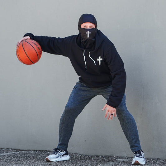 Person wearing a black hoodie with cross designs, blue jeans, and a black mask holding a basketball against a gray wall.