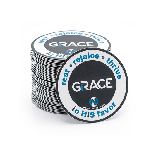 A stack of circular stickers with a dark grey, white, and blue design. The front item reads “GRACE” with smaller text “rest • rejoice • thrive in HIS favor” and a small 'NTL' logo.