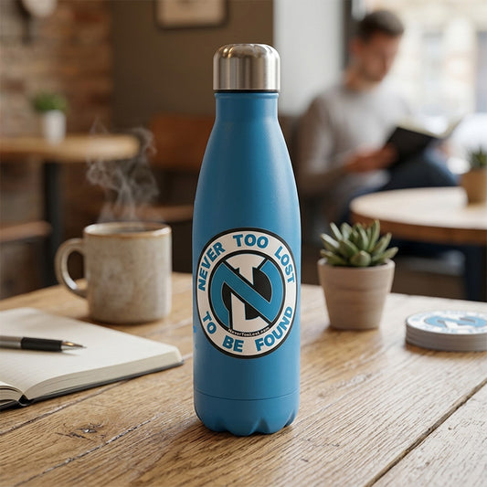 A blue insulated water bottle with a silver cap stands on a wooden table in a cafe setting. The bottle has a circular white, blue, and black graphic on the front that reads 'NEVER TOO LOST TO BE FOUND' with Never Too Lost's 'NTL' compass logo.