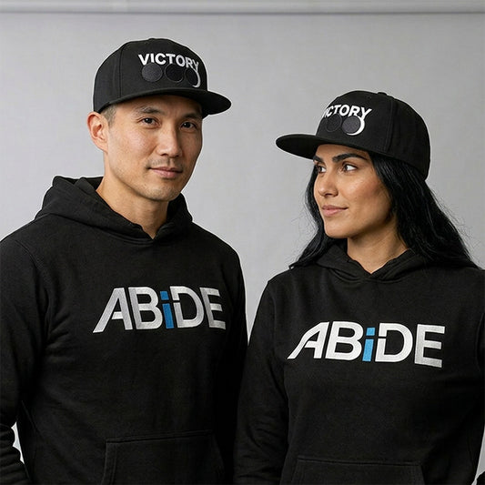 A man and a woman wearing matching black hoodies with a white and blue 'ABiDE' logo on the chest, along with black baseball caps featuring the 'VICTORY' brand logo, posed against a neutral gray background.