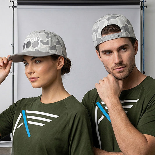 A man and a woman wearing matching olive green "I AM" t-shirts and gray and white camouflage VICTORY in Jesus baseball caps, posed in front of a white studio backdrop.